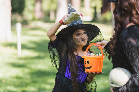 糖果女巫摄影照片_戴着女巫帽的女孩拿着装有糖果的水桶，用玩具手吓着妈妈