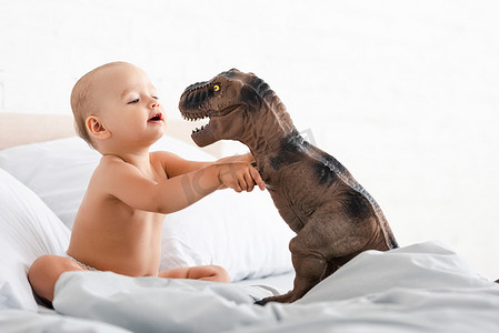 小孩坐在床上，用白毯子，用双手抱着玩具恐龙
