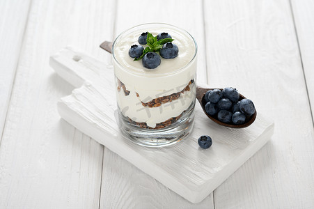 cheese摄影照片_Dessert with cottage cheese, fresh blueberries and granola in a glass on a white wooden background.