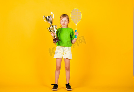 育成摄影照片_快乐的童年小女孩拿着冠军杯和网球拍。羽毛球比赛冠军。胜利和胜利。应该得到的奖励。体育成就。体育成功的概念。孩子们快乐的日子