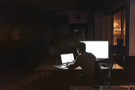 一个年轻的商人坐在办公桌上, 在黑暗的办公室工作, 晚上晚了一台笔记本电脑的倒车