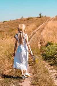 背影走摄影照片_在草地上，穿着白衣、头戴草帽、手持野花走在路上的年轻女子的背影