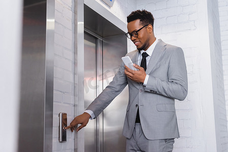 积极的非洲商人在按电梯按钮时使用智能手机