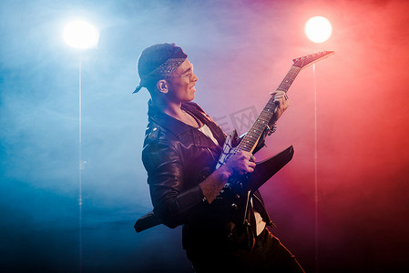 快乐的男性摇滚明星在皮夹克表演在电吉他舞台上与烟雾和戏剧性的照明 