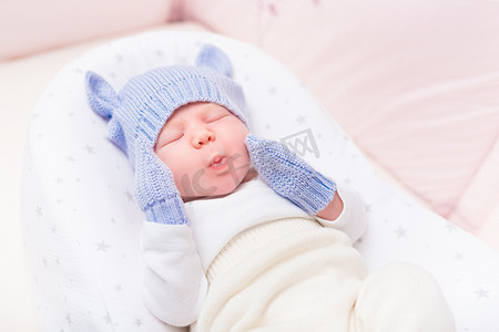 cute摄影照片_Cute little baby wearing knitted blue hat with ears and mittens lying in beautiful cradle with closed eyes and making funny face. Security and childcare concept