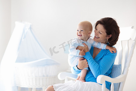 年轻的母亲抱着她刚出生的孩子。妈妈护理宝宝。女人和新出生的男孩，在白色卧室，摇椅和蓝色婴儿床。幼儿园的内部。母亲笑着孩子玩。家人在家里