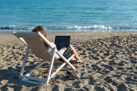 在夏日阳光明媚的日子里,一位穿着办公室衣服的年轻女子带着笔记本电脑在沙滩上。