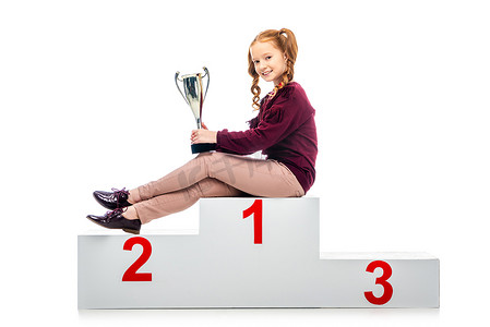 快乐的女学生坐在赢家领奖台上, 拿着奖杯杯, 看着相机孤立的白色