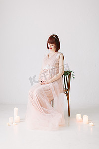 全长的漂亮孕妇在粉红色的礼服坐在装饰椅子上, 在白色地板上燃烧的蜡烛