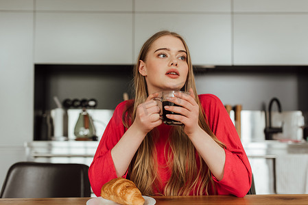 在美味的羊角面包旁边端着一杯咖啡的年轻女人在桌上 