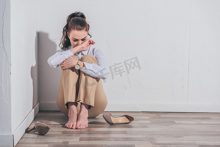悲伤的女人穿着白色的衬衫和米色的裤子坐在地板上, 在家里的墙边哭泣, 悲伤的混乱的概念