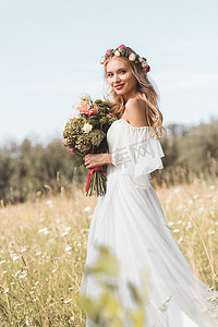 美丽的年轻新娘婚纱礼服和花卉花圈举行花束和微笑的相机