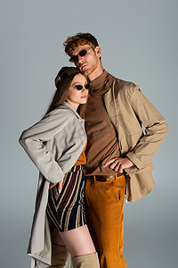 young couple in sunglasses and autumnal clothes posing together isolated on grey