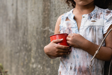 穿着肮脏衣服的可怜的非洲裔美国孩子在城市街道上拿着金属杯的照片 