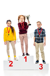 微笑的学龄前孩子与奖牌和奖杯杯子站立在优胜者基座上, 微笑地和看着被隔绝的照相机在白色