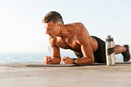 动机 shortless 运动员的耳机做木板演习在一个健身垫在海滩上