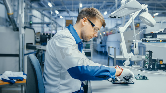 身穿蓝白工作服的年轻人正在使用标语牌组装智能手机的印刷电路板。高科技工厂设施中的电子工厂工人.