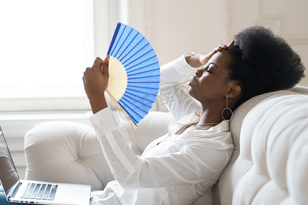 夏季中暑摄影照片_患有中暑的非洲女商人坐在家里客厅里挥动扇子