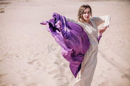 拿着照片摄影照片_一个穿着米色衣服，手里拿着紫色布的年轻纤细姑娘，在沙漠中迎风而坐
