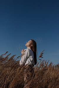 年轻的女人，穿着白衬衫，紧闭双眼，站在蓝天和太阳前干枯的草原上。都市风格和街头时尚。穿休闲装的女孩抬头仰望天空