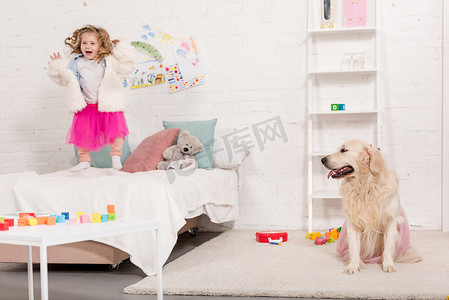 丝质裙子摄影照片_兴奋可爱的孩子跳在床上, 金毛猎犬坐在地毯上粉红色的裙子在儿童房