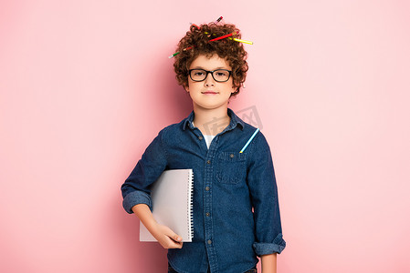 孩子彩色铅笔摄影照片_戴眼镜高兴的孩子，带着彩色铅笔，卷发，手持粉红记事本 