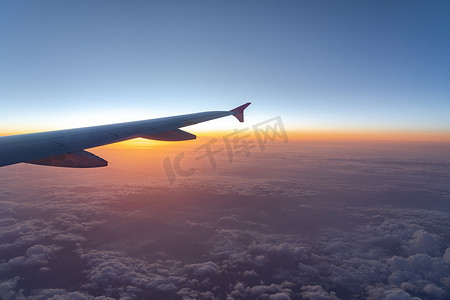 飞机机翼摄影照片_在空中，从飞机窗口看，在太阳升起时，有深蓝色天空地平线和云背景的飞机机翼剪影