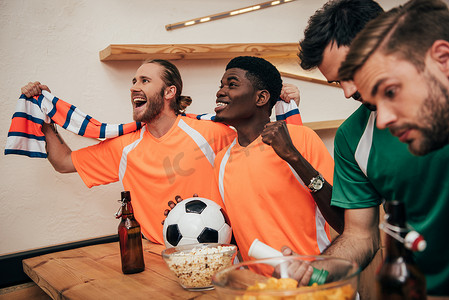 在橙色 t恤和围巾庆祝胜利与球的微笑多文化的球迷, 而他们的不安的朋友在不同的 t恤坐在附近的足球比赛观看在酒吧