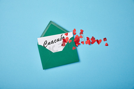 感谢信背景摄影照片_绿色信封与感谢信在白色卡片上用俄语和红色纸心在蓝色背景