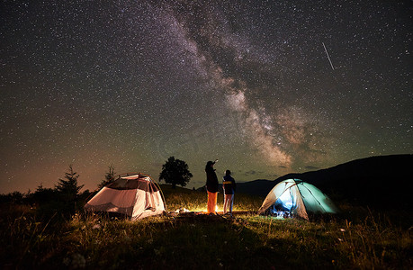 篝火摄影照片_后视图的母亲和儿子的游客休息在山上露营, 站在篝火旁和两个帐篷, 看着夜空中充满了星星和银河, 享受夜晚的景象。女人指着天空