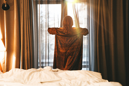 遇见胡旋摄影照片_一个女人在卧室的窗前遇见了黎明，她穿着丝绸长袍拉开窗帘