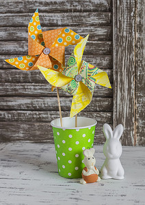 Homemade paper pinwheel, ceramic bunnies on a light rustic wood table. Easter still life in vintage style