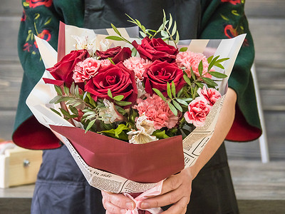 花束包装摄影照片_在花匠的手里，有一束红玫瑰、桉树和康乃馨。包装上的一束花。围裙上的花匠，臂膀上的花束。花店的花匠