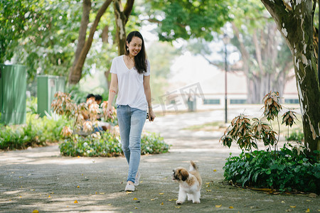 泛亚洲摄影照片_一个年轻的泛亚洲妇女的肖像坐在她年轻的石 tzuh 小狗在公园里的一天。她在微笑, 他们都在享受温暖的一天。.