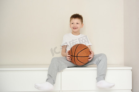 可爱的笑脸摄影照片_穿着休闲装的可爱的笑脸男孩坐在房间的梳妆台上，手里拿着篮球。白色背景。案文的篇幅