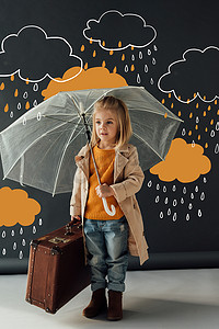 黑色皮箱摄影照片_穿着风衣和牛仔裤的孩子在黑色背景的幻想雨下拿着雨伞和皮箱