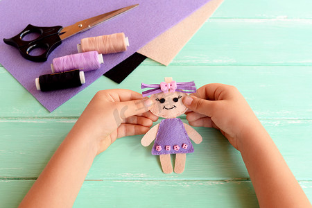 手工纸底纹摄影照片_孩子他手里拿着一个感觉的娃娃，并显示它。剪刀，线程，觉得桌上的床单。可爱的毛绒的玩具是由手工做成。孩子容易缝纫项目 