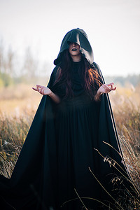 一个身穿黑色连衣裙的女孩，披着斗篷的斗篷，站在森林背景下的高干草地上。女巫服，撒旦师，亡灵法师，万圣节服装.