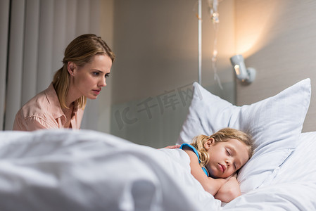 看病床摄影照片_担心的母亲看着生病的孩子睡在病床上的医院