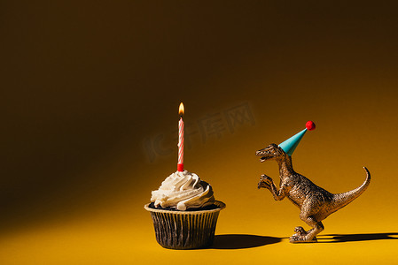 茶杯蛋糕摄影照片_戴着派对帽的玩具恐龙，旁边是茶杯蛋糕，背景为褐色，点着蜡烛