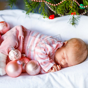 一个星期大的新生婴儿，在圣诞树附近带着粉色球，背景上有五彩缤纷的花环灯。 可爱的孩子的衣服，小女孩睡觉。 家庭，圣诞节，出生，新生活