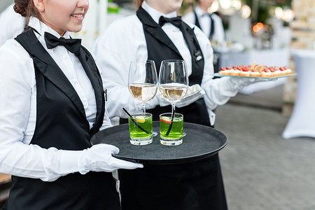 一杯酒卡通摄影照片_在自助餐宴会、节庆活动或婚礼期间，有一半的职业服务员身穿制服，为客人提供葡萄酒、鸡尾酒和零食。满满一杯酒放在盘子里.户外派对饮食服务