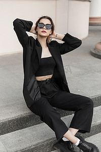 Trendy fashionable young beautiful woman model with modern sunglasses in elegant business urban outfit with top, blazer, pants and shoes sits on the steps on the street