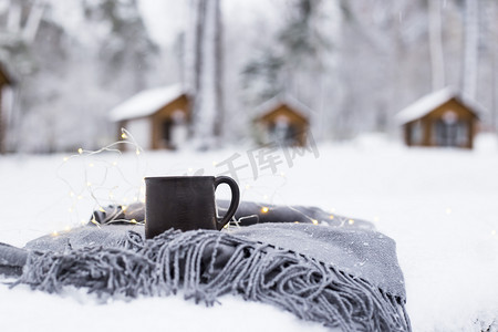 一杯热咖啡在一条灰色的围巾上, 在一个冬天森林里的木制房子旁边的白雪覆盖的桌子上