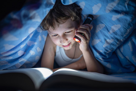 对的人摄影照片_快乐的男孩躺在毯子下，用手电筒在黑暗中看书，而不是在每个人都睡着的时候睡觉。那孩子躲得远远的对知识的渴望.