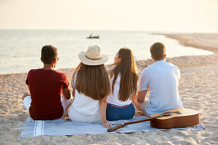 在度假期间，一群朋友坐在白色沙滩上的毛巾上，享受海上的日落。男人和女人在岸上用吉他放松。宁静场景.