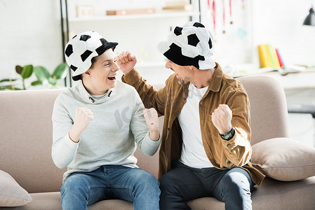 兴奋的父亲和十几岁的儿子在足球帽看比赛, 在家里的沙发上尖叫, 看着对方