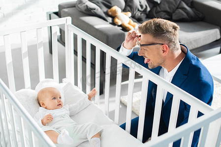 幸福的父亲在正式的服装显示舌头对幼女在婴儿床 