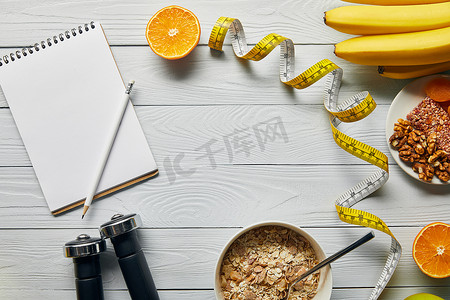 测量磁带，哑铃，饮食食品和木制白色背景的空白笔记本的顶视图 