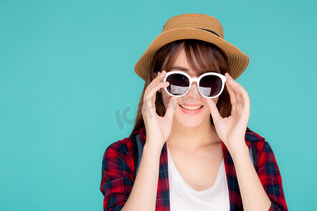 美丽的肖像画年轻的亚洲女人头戴帽子，戴着太阳镜，面带微笑，充满自信地享受在蓝色背景下度过的暑假之旅，旅游的亚洲女孩带着旅游的快乐.